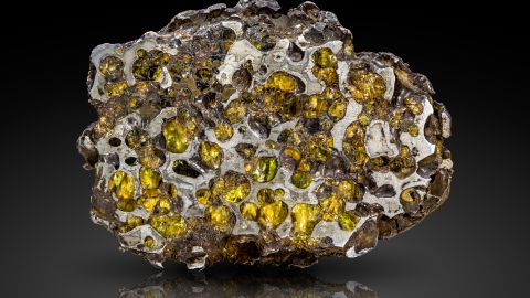 Fénykép a Pallasit meteoritról, a csiszolt felületen a zöldessárgán ragyogó olivin kristályokat körbeöleli a fémesen csillogó vas